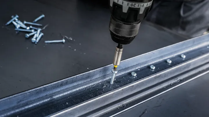 Power drill driving a screw into a metal beam on a workbench.