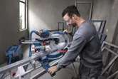 A person wearing safety equipment operates a mitre saw to cut a metal piece.