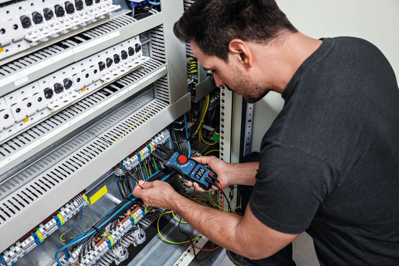A person tests electrical wires in a control panel using a fork meter.