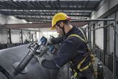 A person wearing safety equipment uses an impact wrench to tighten bolts on a large metal tank.