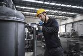 A person wearing safety equipment uses a cordless impact wrench on a large metal pipe.