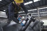 A person wearing safety equipment uses a cordless impact wrench on industrial piping.