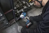 A person wearing safety equipment tightens a bolt on industrial machinery with a cordless impact wrench.