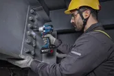 A person wearing safety equipment uses a cordless impact wrench on a steel beam.