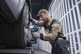 A person wearing safety equipment uses a cordless impact wrench on a car wheel.