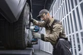 A person wearing safety equipment uses a cordless impact wrench on a car wheel.
