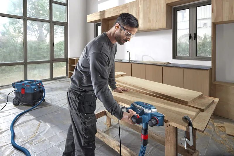 GHO 16-82 A person wearing safety equipment uses a planer on wooden boards in a workshop.