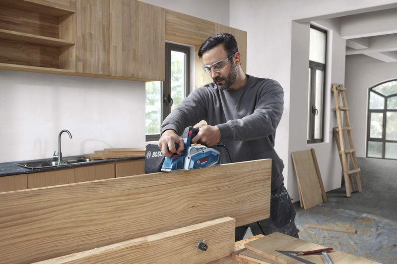 GHO 16-82 A person wearing safety equipment uses a planer to smooth a wooden board in a workshop.