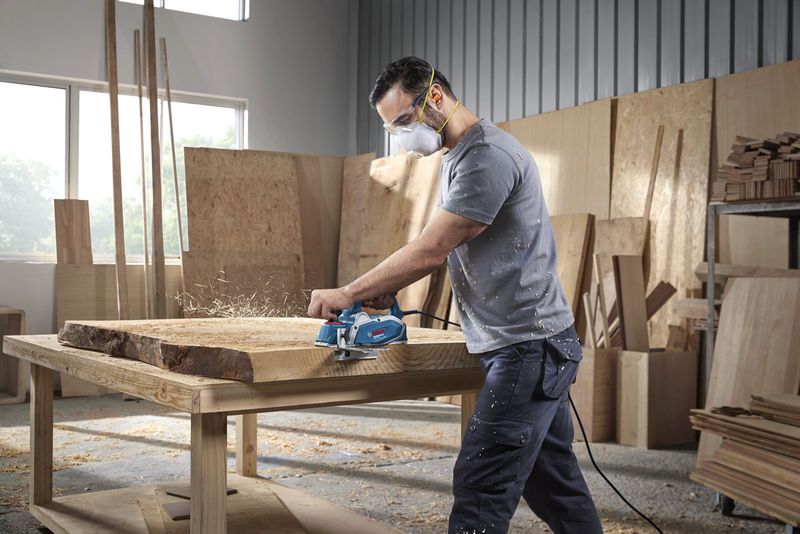 A person wearing safety equipment planes a thick wooden slab in a carpentry workshop.