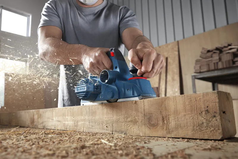 GHO 16-82 A person wearing safety equipment planes a wooden beam, wood shavings flying.