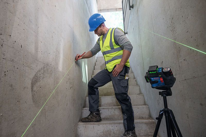 A person wearing safety equipment marks a wall using a laser leveling tool on a tripod.