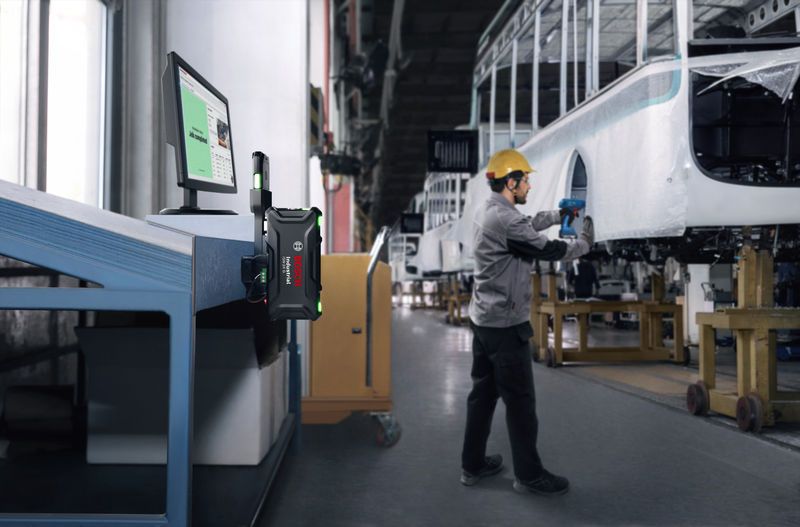 A person wearing safety equipment uses a power tool to assemble a vehicle frame in a factory.