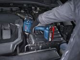 A person wearing safety equipment uses a cordless impact driver on a car engine.
