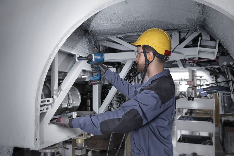 A person wearing safety equipment uses an impact wrench on a metal frame.