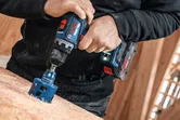 A person drills a large hole into wood using a power tool in a workshop.
