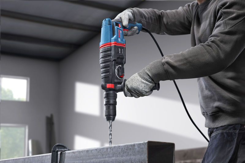 A person wearing safety equipment drills into a metal beam using a rotary hammer.