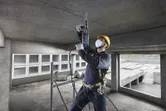 A person wearing safety equipment drills into a concrete ceiling with a rotary hammer.
