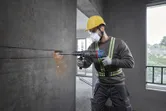 A person wearing safety equipment uses a rotary hammer to drill into a concrete wall.
