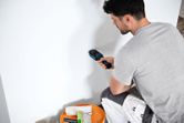 A person uses a material moisture meter against a white wall.