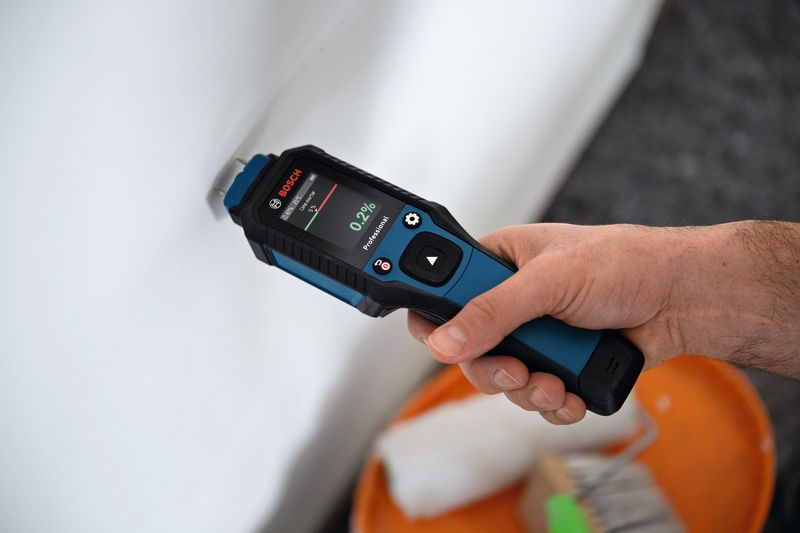 A person uses a material moisture meter to check a white wall.