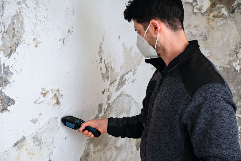 A person wearing safety equipment uses a material moisture meter on a damaged wall.