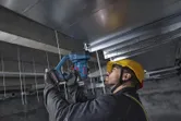 A person wearing safety equipment uses a cordless threaded rod cutter on ceiling rods.
