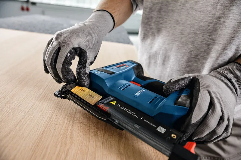 A person wearing safety equipment loads nails into a cordless nail gun on a wooden surface.