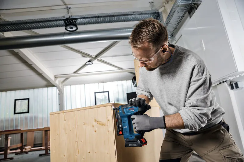 A person wearing safety equipment staples wood panels together in a workshop.