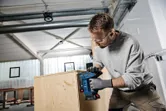 A person wearing safety equipment staples wood panels together in a workshop.