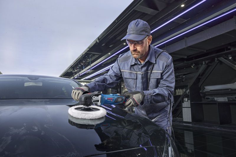 A person wearing safety equipment polishes a black car with a power polisher.