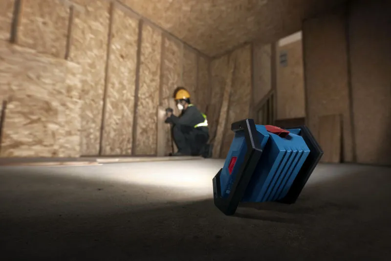 A person wearing safety equipment installs drywall, illuminated by a cordless jobsite light.