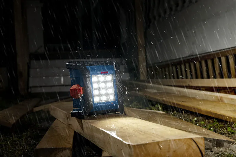 Cordless jobsite light illuminating wooden planks in heavy rain at a construction site.