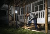A person wearing safety equipment drills wood on a porch, illuminated by a cordless jobsite light.