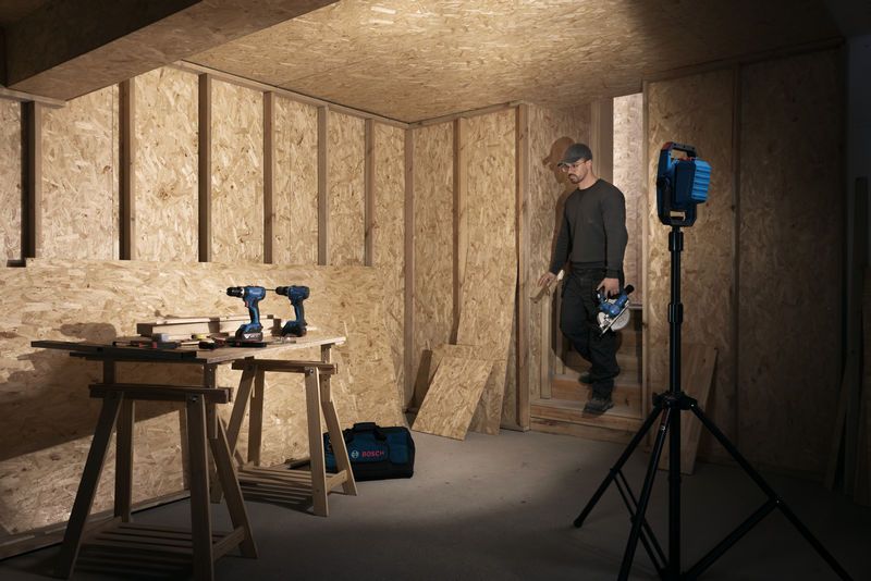 A person wearing safety equipment walks into a wood workshop lit by a cordless jobsite light.