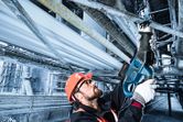 A person wearing safety equipment uses a reciprocating saw to cut a metal pipe overhead.