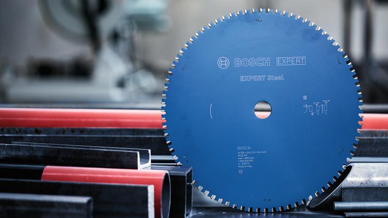 EXPERT Steel Circular Saw Blade, 305 x 2.6/2.2 x 25.4 mm, T60 Circular saw blade positioned upright beside metal and plastic pipes in a workshop.