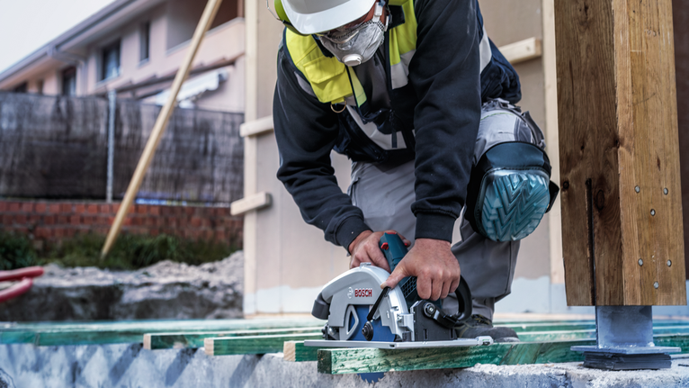 Person wearing safety equipment cuts wood with a circular saw on a construction site.