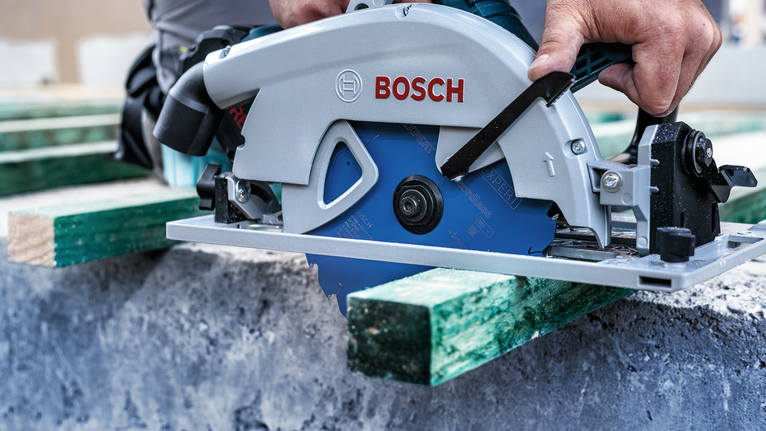 Person operates a circular saw to cut a green wooden beam on a construction site.