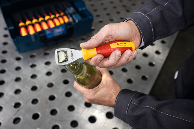 Person in workwear uses a screwdriver handle to open a bottle on a metal workbench.
