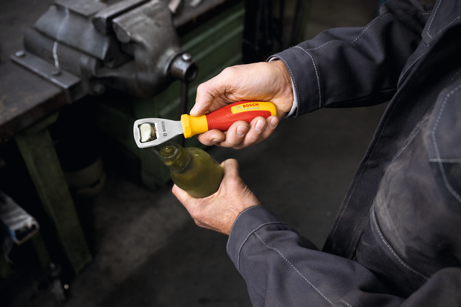 Person in workwear opens a bottle with a screwdriver in a workshop.