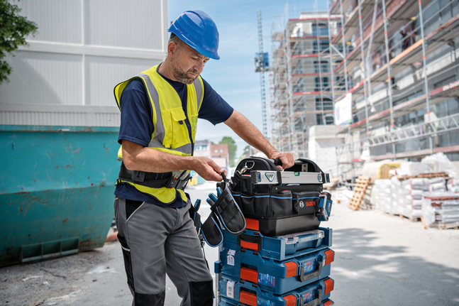 A person wearing safety equipment organizes a tool bag on stacked toolboxes at a construction site.