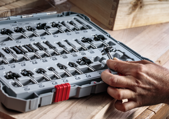 Person selects a router bit from an organized tool set on a wooden workbench.