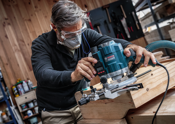 Person wearing safety equipment uses a router to shape the edge of a wooden board.