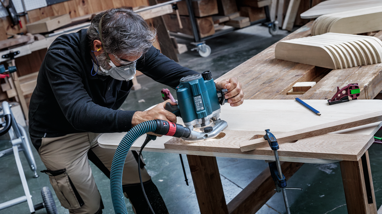 Person wearing safety equipment shapes wood using a power router in a workshop.