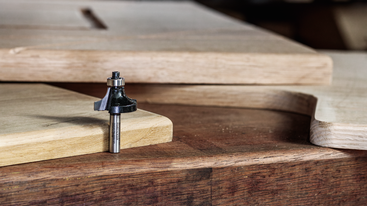 Router bit resting on a wooden workbench beside cut and shaped wood panels.