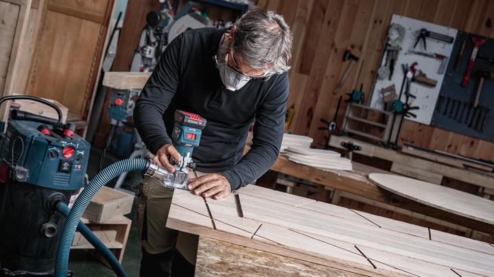 Person wearing safety equipment uses a power tool to rout grooves into a wooden board.