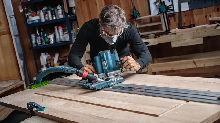 Person wearing safety equipment guides a router along a track to shape a wooden board.