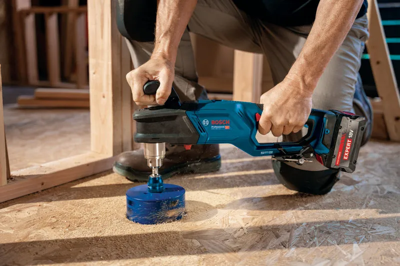 A person uses a cordless angle drill to bore a large hole in a wooden floor.