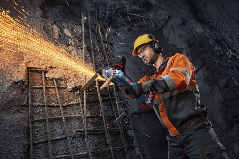 GWS18V-11PS Worker wearing safety equipment grinds rebar in concrete, creating sparks.