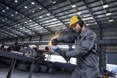 A person wearing safety equipment grinds a metal pipe, producing sparks in a factory.